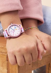 Rosa Uhr mit floralem Design auf einem weißen Ziffernblatt, Textilarmband in Pastellfarben. Armband mit mehrfarbigen Perlen am Handgelenk.