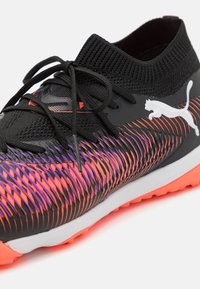 Zwarte sportieve schoen met een gestructureerde bovenkant met een paarse en oranje patroonverloop, elastische manchet en een opvallend wit logo.