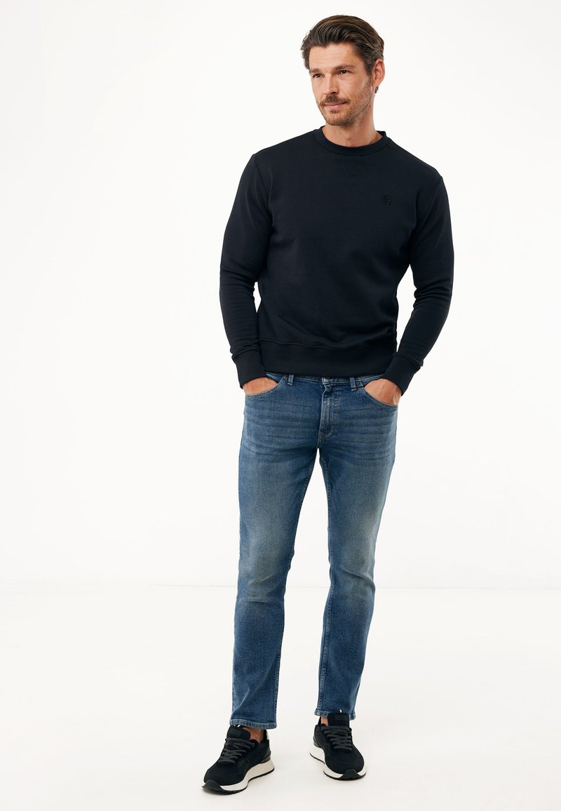 Czarna bluza z ściągaczami na rękawach, w połączeniu z jasnoniebieskimi dżinsami. Buty są czarne z białą podeszwą. Całość tworzy casualowy wygląd.