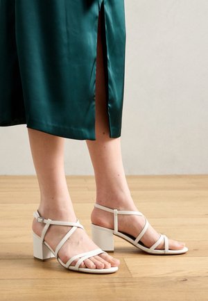 Femme portant des sandales à talons blocs blancs avec des brides et une jupe satinée teal avec une fente latérale sur un sol en bois.