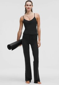 Juodas sportinis tanktopas su plonomis petnešomis derinamas su juodomis kelnėmis, turinčiomis kaspinį. Modelis laiko suvyniotą jogos kilimėlį.