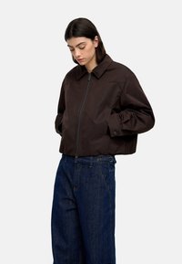 Brązowa krótka kurtka z wysokim kołnierzem i przednim zamkiem, w połączeniu z luźnymi niebieskimi dżinsami. Kurtka ma guziki na rękawach.