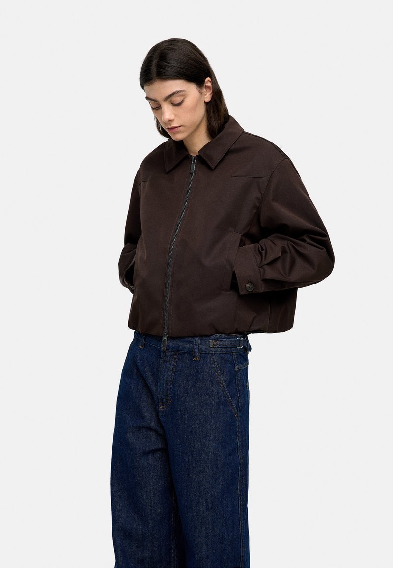 Brązowa krótka kurtka z wysokim kołnierzem i przednim zamkiem, w połączeniu z luźnymi niebieskimi dżinsami. Kurtka ma guziki na rękawach.