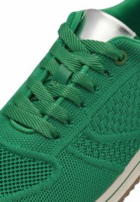 Botte de sport verte avec une tige en maille texturée, des lacets plats et un accent argenté au niveau du talon. Semelle en caoutchouc avec un motif de traction marron.