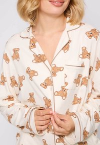 Pajama top in white fabric featuring brown teddy bear prints. Design includes a chest pocket, button closure, and contrasting piping.