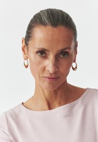 Ritratto di una donna con capelli grigi, che indossa orecchini a cerchio dorati e una maglietta rosa pallido con scollatura ampia, tessuto morbido e cuciture sottili.