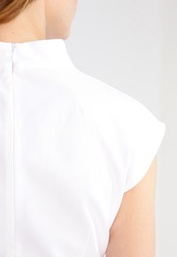 Prenda blanca texturizada con un cuello alto y mangas cortas con un acabado; presenta un discreto cierre en la parte trasera.