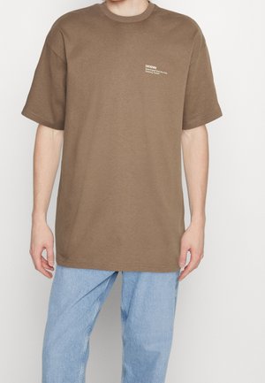 Osoba ubrana w brązowy oversize T-shirt z małym białym napisem na klatce piersiowej i jasnoniebieskie luźne dżinsy, stojąca na białym tle.