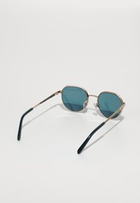 Occhiali da sole rotondi con lenti teal, montatura in metallo dorato e templi verde scuro. Presentano morbidi cuscinetti per il naso e un design elegante.