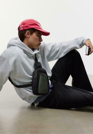 Borsa a tracolla nera e verde oliva con una finitura testurizzata, indossata da una persona in felpa grigia e jeans scuri, seduta su uno sfondo neutro.