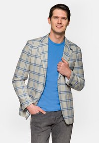 Jasnobeżowa marynarka w niebieską kratę, połączona z niebieską koszulką i szarymi dżinsami. Marynarka ma wąskie klapy i przednie kieszenie.