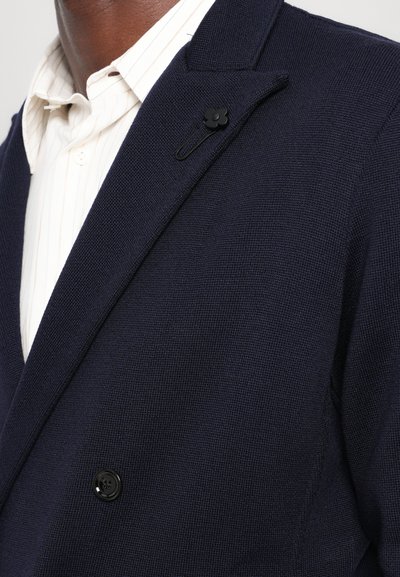 Navy blazer featuring a textured fabric, notched lapel, and a single black button accent. Worn over a light striped shirt.