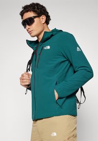 Chaqueta con capucha color verde azulado, con un acabado texturizado, bolsillos con cremallera y cremalleras negras. Lleva unas gafas de sol y pantalones beiges.