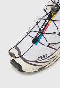 Scarpa sportiva bianca con una parte superiore in rete testurizzata, lacci neri e colorati, marchio in evidenza e una spessa suola in gomma nera.