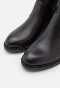 Bottes noires en cuir à hauteur de cheville avec une finition polie, bout pointu et panneaux latéraux élastiques. Présentent une couture autour de la semelle.