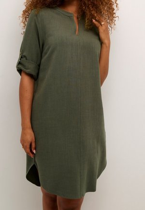 Mujer con vestido suelto de color verde oliva hasta la rodilla, con mangas remangadas y un pequeño escote en V, de pie contra un fondo liso.