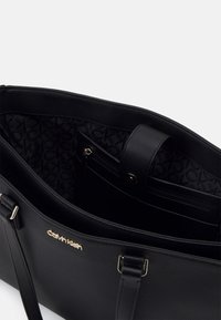 Czarna skórzana torba tote z złotym logo. Wnętrze zawiera kieszeń na zamek i wzorzystą podszewkę z logo CK. Wytrzymałe podwójne uchwyty do noszenia.