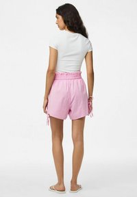 Rožiniai čekiniai šortai su raišteliu juosmens, šoniniais mazgais ir laisvu siluetu. Modelis vilki baltą prigludusią marškinėlius ir šlepetes.