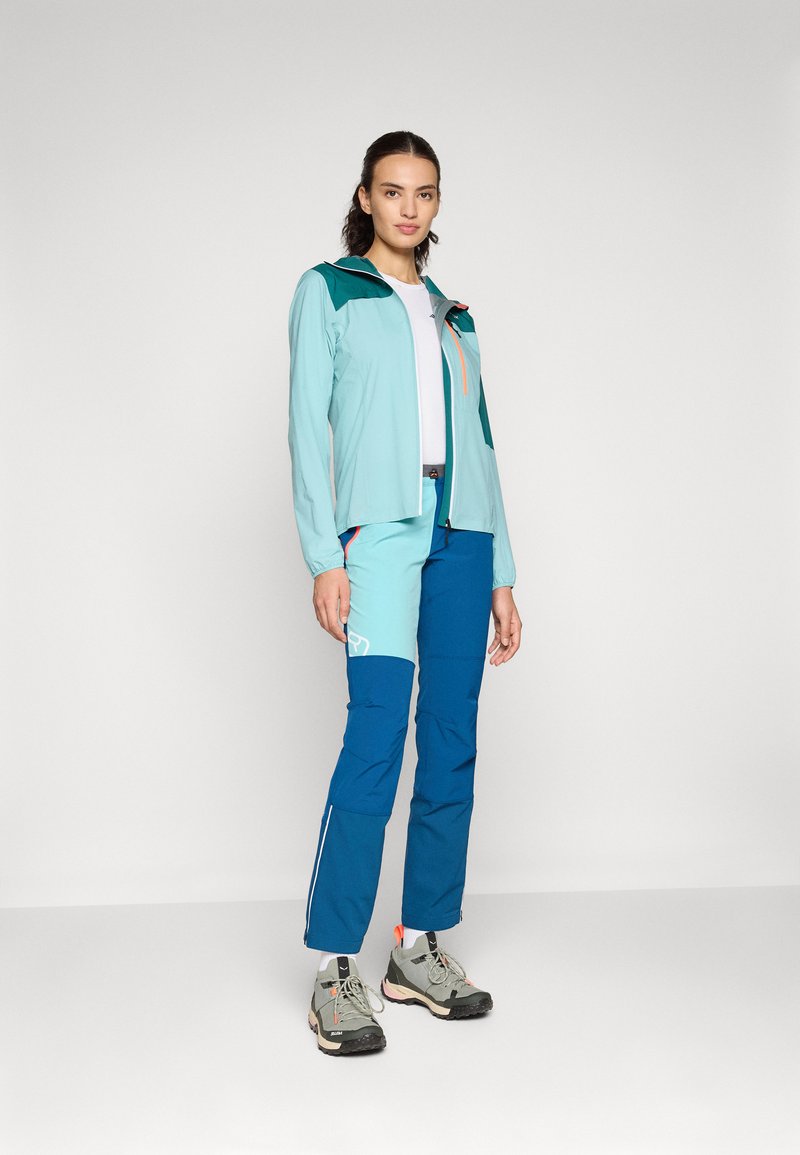 Helles blaues Jackett mit dunkelblauen Akzenten und orangefarbenem Reißverschluss, kombiniert mit blauen Hosen mit Taschen und weißen Streifen. Graue Wanderschuhe.