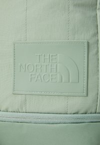 Tela verde claro con parche de logo en relieve; textura suave y detalles cosidos cerca de la parte inferior, con cierre de cremallera.