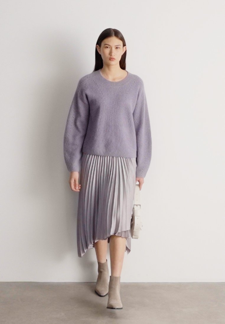 Femme marchant, portant un pull en tricot lavande, une jupe midi plissée argentée, des bottines beige, tenant un sac à main blanc.