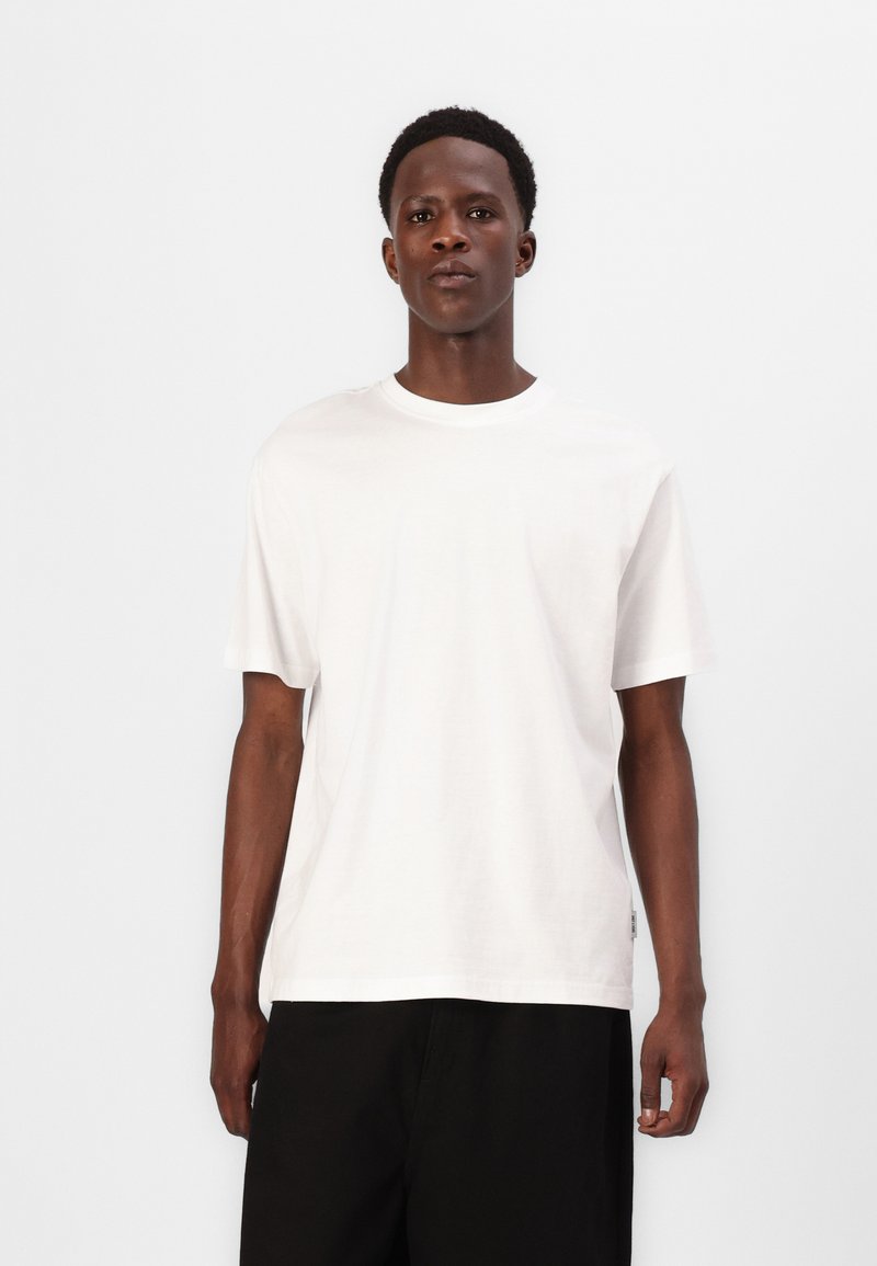 Weißes Baumwoll-T-Shirt mit Rundhalsausschnitt und kurzen Ärmeln, mit lockerer Passform. Das Modell trägt es zu schwarzen Shorts.