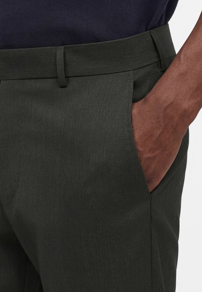 Σκούροι ελαιδί παντελόνια φορεμένα με το χέρι εν μέρει στην πλαϊνή τσέπη και μια εμφανή θηλιά ζώνης στη μέση.