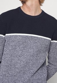 Pull en maille bleu marine foncé et gris clair présentant un motif rayé texturé avec une ligne accentuée blanche traversant la poitrine et un col rond.