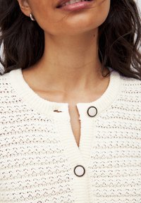 Gilet léger de couleur crème avec un design en tricot texturé et ouvert, présentant un col rond et deux boutons contrastants à l'avant.