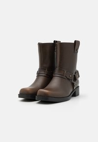 Bottines en cuir marron, avec une texture lisse, des sangles sur les côtés, des éléments en métal et un petit talon bloc. Le design est épuré et pratique.