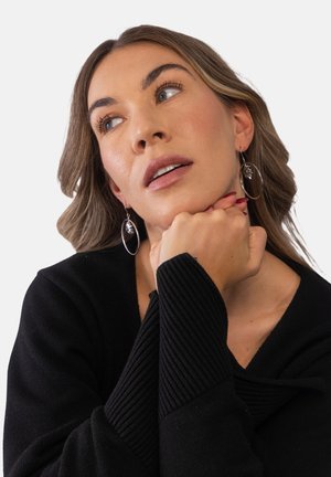 Woman with shoulder-length light brown hair wearing black sweater and dangling leaf earrings, resting chin on clasped hands, looking upward.