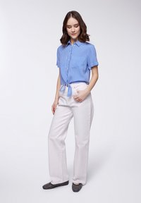 Camicia a righe blu con bottoni e maniche corte, annodata in vita. Pantaloni bianchi a gamba larga e scarpe nere con borchie. Texture morbide.