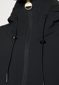 Černá nepromokavá bunda s vysokým límcem, zlatým jezdcem zipu a nastavitelnými stahovacími šňůrkami. Má hladký, texturovaný povrch.