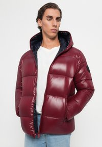 Young man wearing a burgundy puffer jacket with a hood over a white shirt and light blue jeans, standing against a plain backdrop.