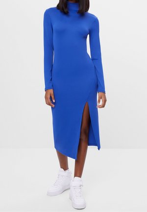 Woman wearing a long-sleeve royal blue midi dress with high slit, paired with white high-top sneakers, against a plain background.