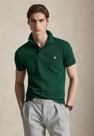 Jeune homme portant un polo vert foncé et un pantalon gris clair rayé, mains dans les poches, sur un fond uni.