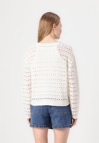 White knitted sweater with openwork pattern and ribbed cuffs, paired with denim shorts. The sweater has a relaxed, slightly cropped fit.