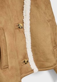 Giacca scamosciata marrone chiaro con interno in sherpa bianco, due bottoni in tartaruga e tasche laterali, visibile in primo piano.