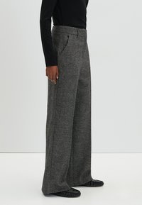 Wijde pantalon in een grijze, gestructureerde stof met een op maat gemaakte tailleband en zijzakken, gecombineerd met een zwart aansluitend shirt met lange mouwen en platte schoenen.