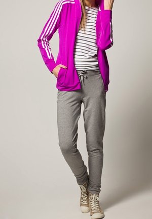 Giacca sportiva fucsia con strisce bianche, abbinata a pantaloni da tuta grigi. Sneakers alte beige completano l'outfit.