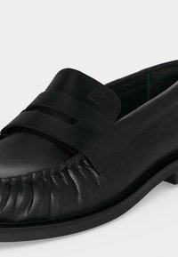 Mocassin en cuir noir avec coutures moc toe, bride penny et logo embossé sur la languette, présenté sur fond blanc.