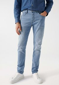 Svijetloplave traperice imaju uski kroj, pet džepova, rajferšlus i blago izblijedjelu teksturu, uparenih s bijelim tenisicama.