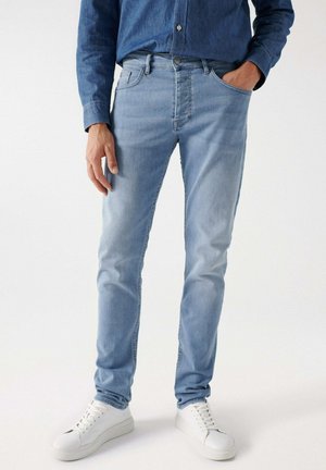Los jeans de mezclilla azul claro tienen un ajuste slim, cinco bolsillos, un cierre con cremallera y una textura ligeramente desvanecida, combinados con zapatillas blancas.