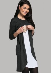Schwarze leichte Strickjacke über einem weißen Kleid, mit drapiertem Design und 3/4-Ärmeln. Sichtbares verstellbares Multifunktionspaneel.