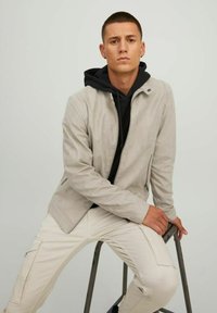 Light gray suede jacket with a zip front, a standing collar, and side pockets, worn over a black hoodie and paired with cream-colored trousers.