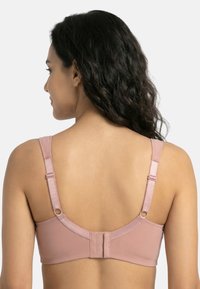 Kobieta z długimi kręconymi włosami odwrócona tyłem, ubrana w jasno-różowy regulowany biustonosz z zapięciem na haftki na jednolitym tle.