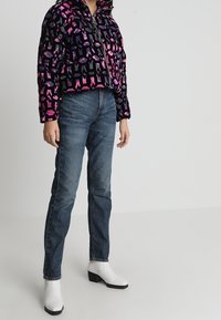 Korte zwarte jas met een geometrisch roze en paarse patroon, gecombineerd met blauwe jeans en witte enkelboots met een blokhak.
