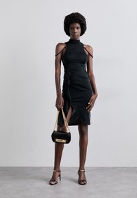 Vestido negro ajustado con cortes en los hombros, detalle fruncido y abertura alta en el muslo. Combinado con un bolso negro de Moschino y sandalias de tacón negras.