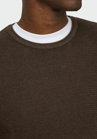 Pull brun texturé avec un motif en chevrons, doté d'un col rond. En dessous, une chemise blanche à col est visible.