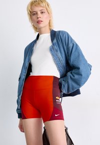 Denim bomber jacketas ant balta prigludusia marškinėliais, derinamas su aukšto liemens raudonais ir vyno spalvos sportiniais šortais, išsiskiriančiais Nike logotipu ir grafika.